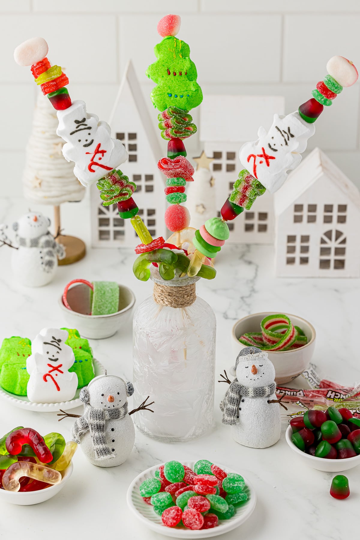Skewers with assorted gummy Christmas candies on them, a white christmas village in the background. 