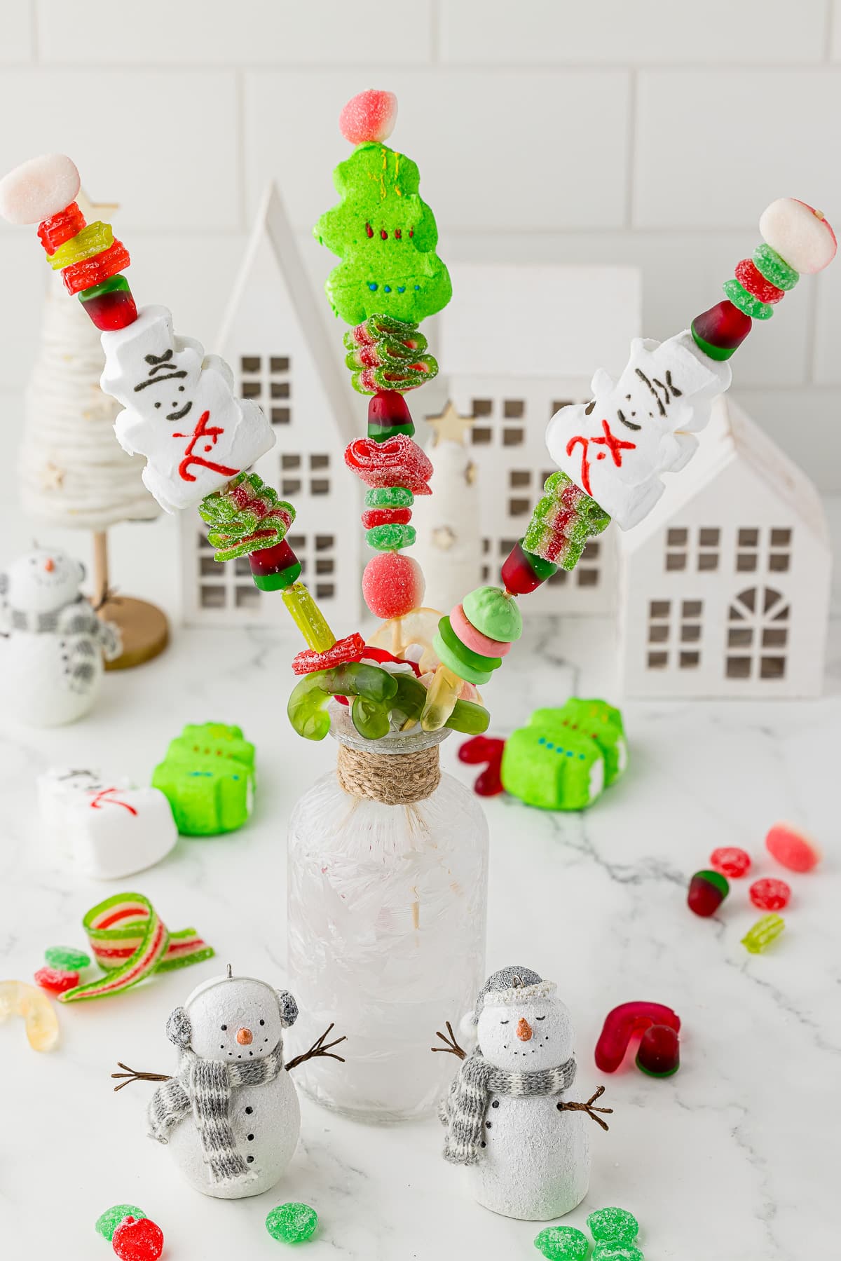 Scattered christmas gummy candy on a countertop with a white christmas village in the background and three candy kabobs in a jar.