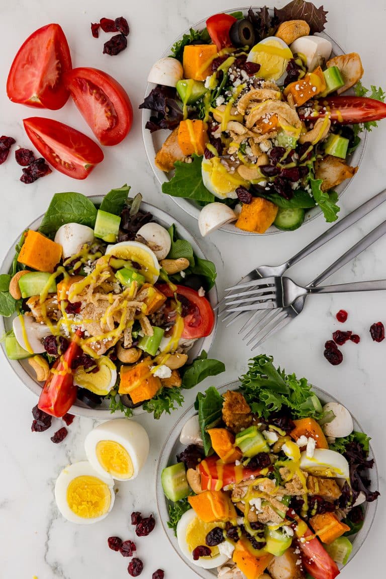 3 white plates filled with salad that include eggs, craberries, roasted squash, cucumbers, tomatoes, and dressing with mushrooms and lettuce