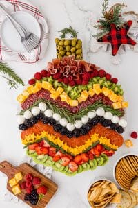 charcuterie board made to look like a round Christmas ornament with meats, fruits, cheese, and sprigs of rosemary on a white marble countertop, with small white plates, 3 forks, and bowls of crackers.