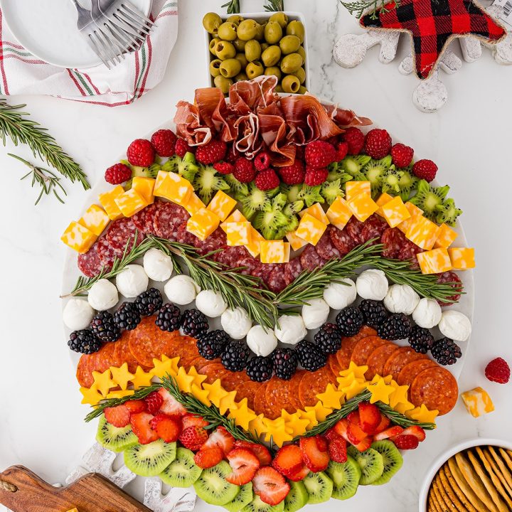 charcuterie board made to look like a round Christmas ornament with meats, fruits, cheese, and sprigs of rosemary on a white marble countertop, with small white plates, 3 forks, and bowls of crackers.