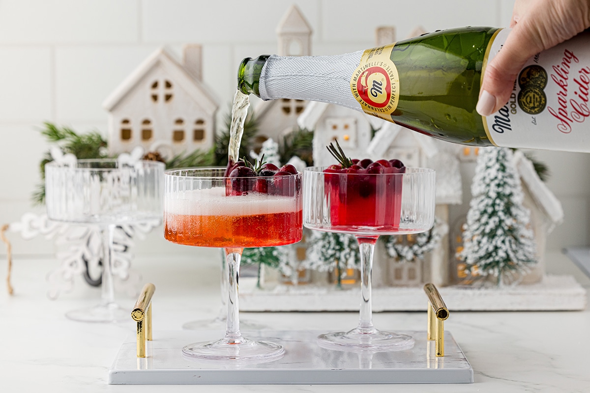pouring martinelli's sparkling cider over a square ice cube filled with cranberries in two coupe glasses, with one empty coupe glass in the background with a white winter villate