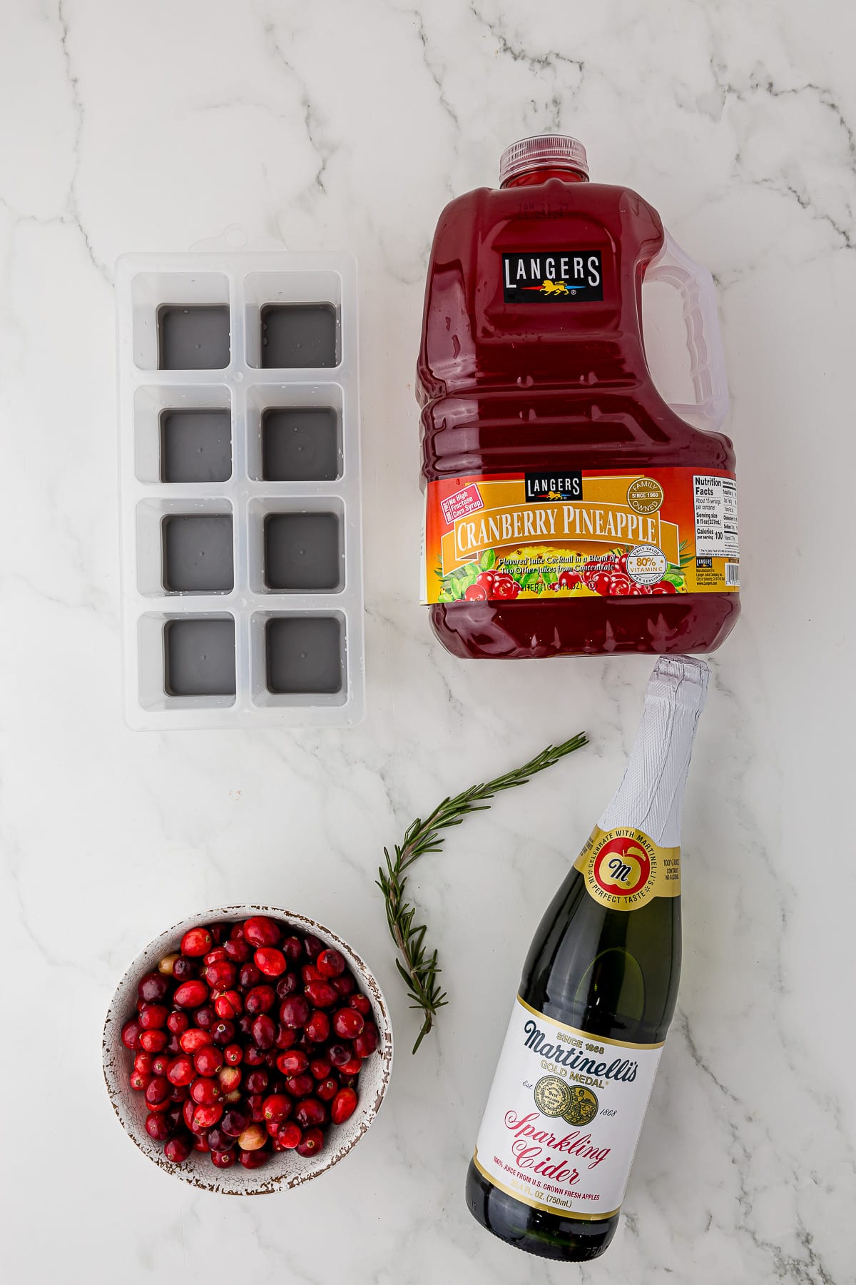square ice cube tray, langer's cranberry pineapple juice, a sprig of rosemary, a bowl of cranberries, and a bottle of martinelli's sparkling cider all on a white countertop