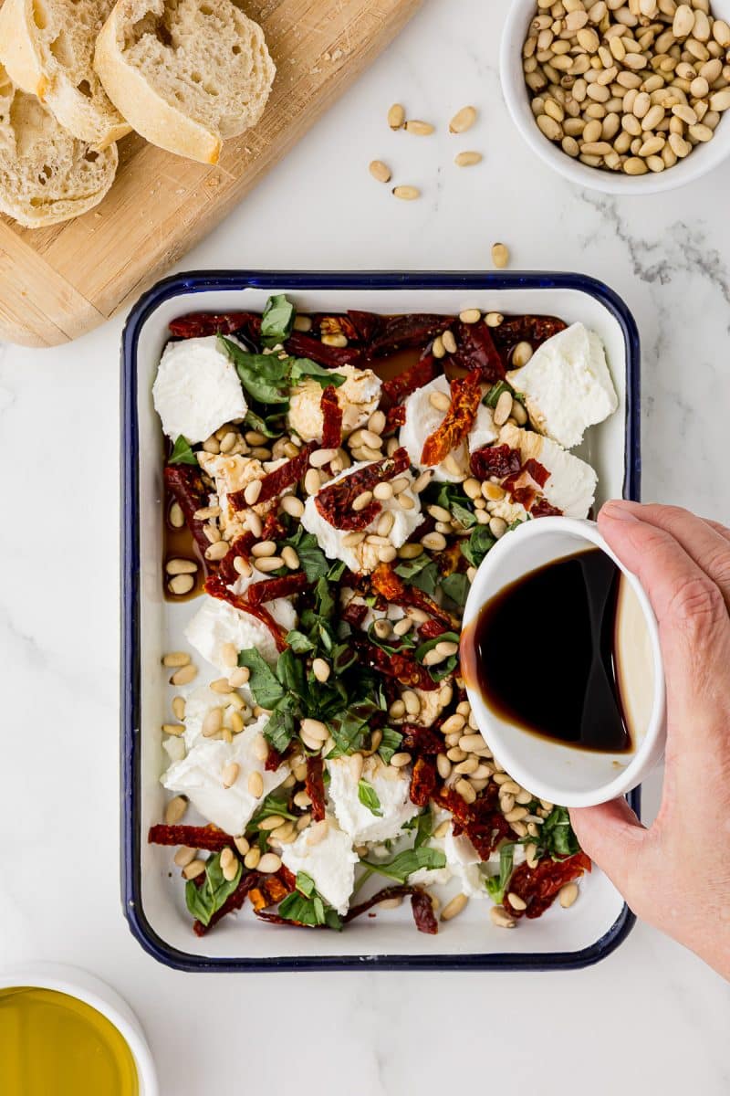 pouring balsamic vinegar over goat cheese, sun-dried tomatoes, pine nuts, and basil in a white serving dish