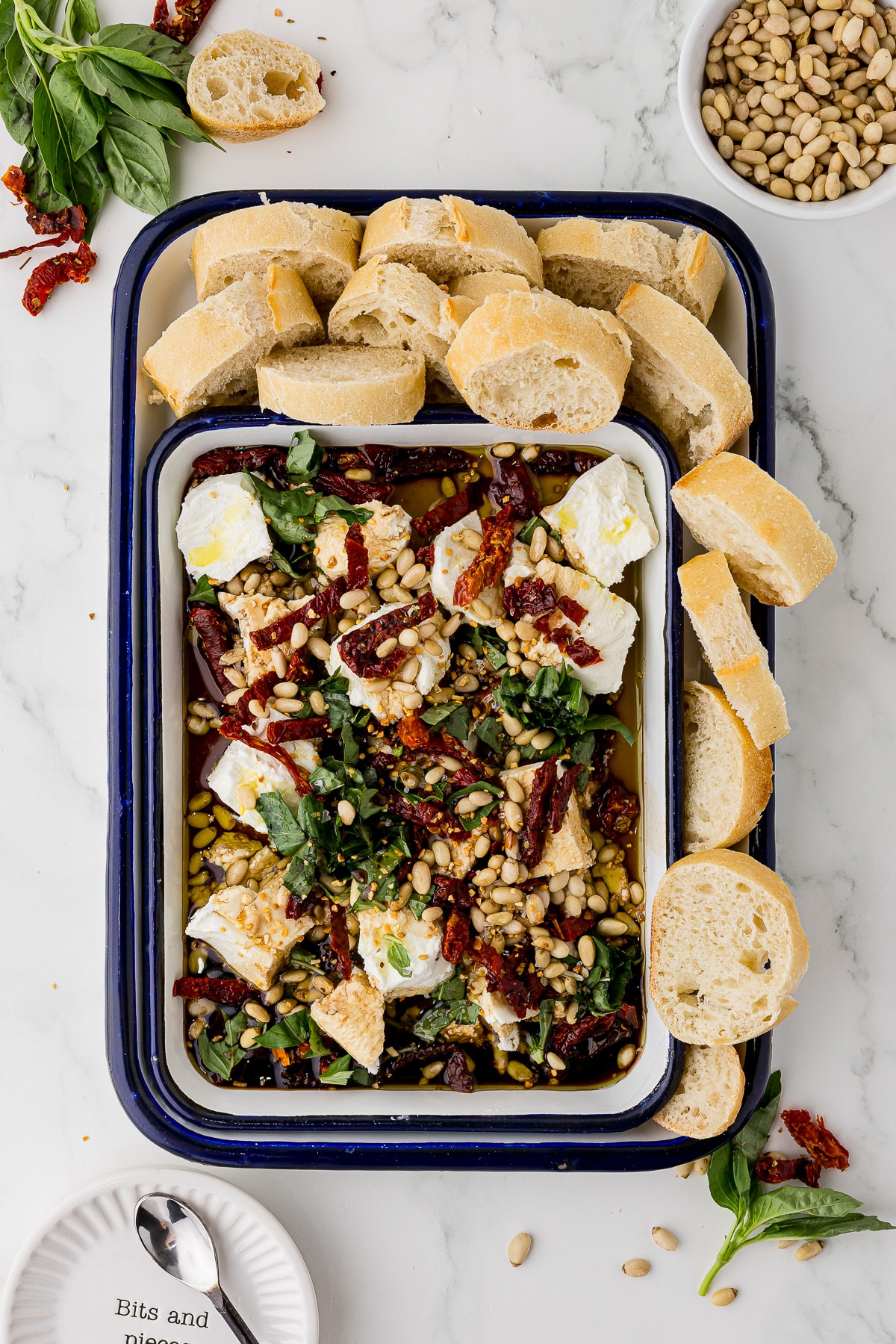 goat cheese appetizer in a white serving dish, surrounded by bread slices on a white marble countertop, a bowl of pine nuts and scraps surrounding the dishes.
