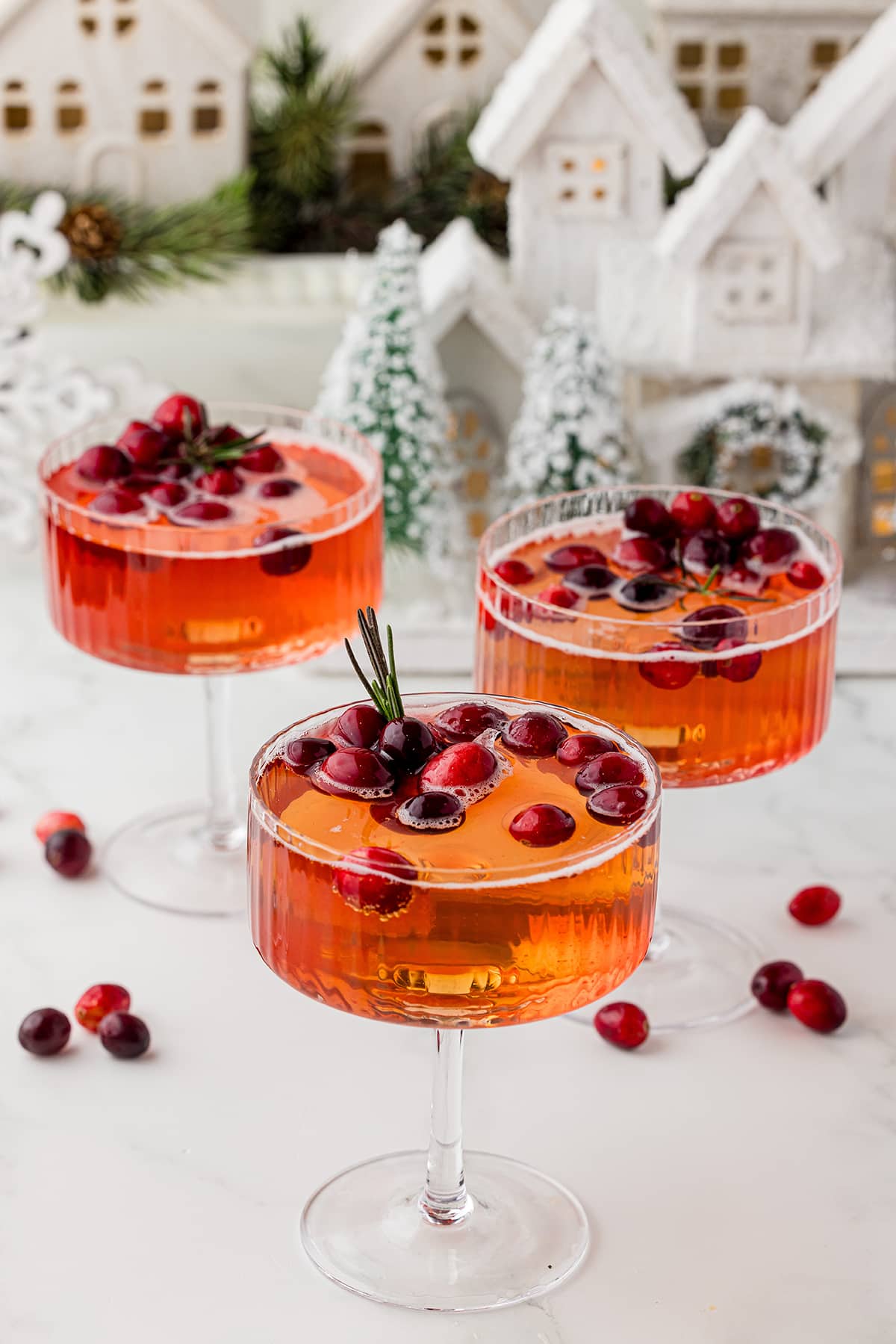 three coupe glasses with floating cranberries and a sprig of rosemary on a white countertop with cranberries on the counter, a white winter village in the background