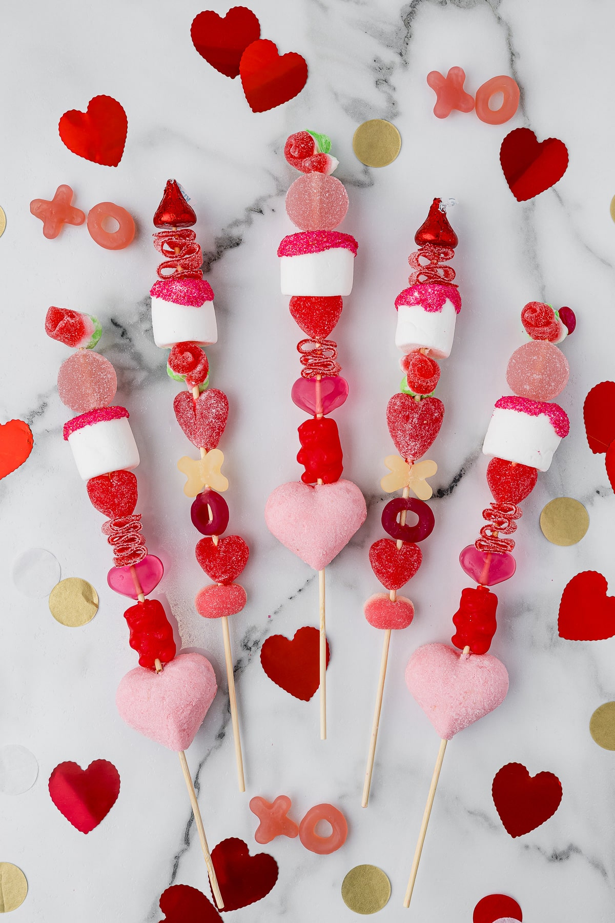5 valentine's day candy kabobs on a white marble countertop with red foil hearts and gummy x's and o's