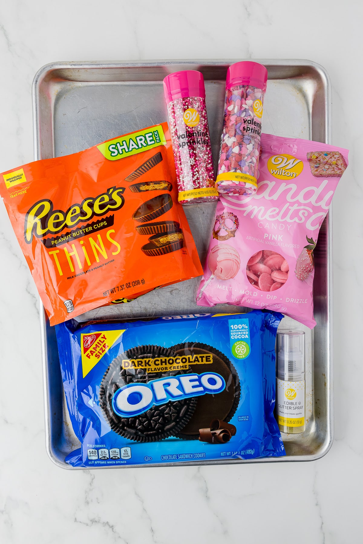 a package of Reese's peanut butter things, wilton sprinkles, wilton pink candy melts, dark chocolate flavor creme oreos, and edible glitter spray on a cookie sheet