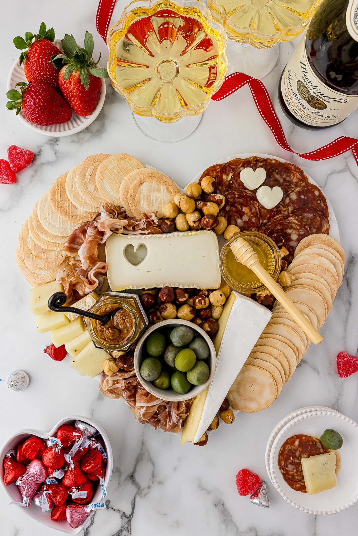 Meats and cheeses spread out on a heart shaped cutting board with drinks and fruit surrounding the tray