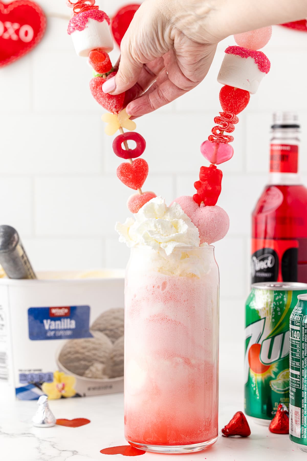 clear glass with one scoop of ice cream on a white counter with vanilla ice cream, canned whipping cream, strawberry syrup, and a bottle of 7-up on a white counter with two candy kabobs