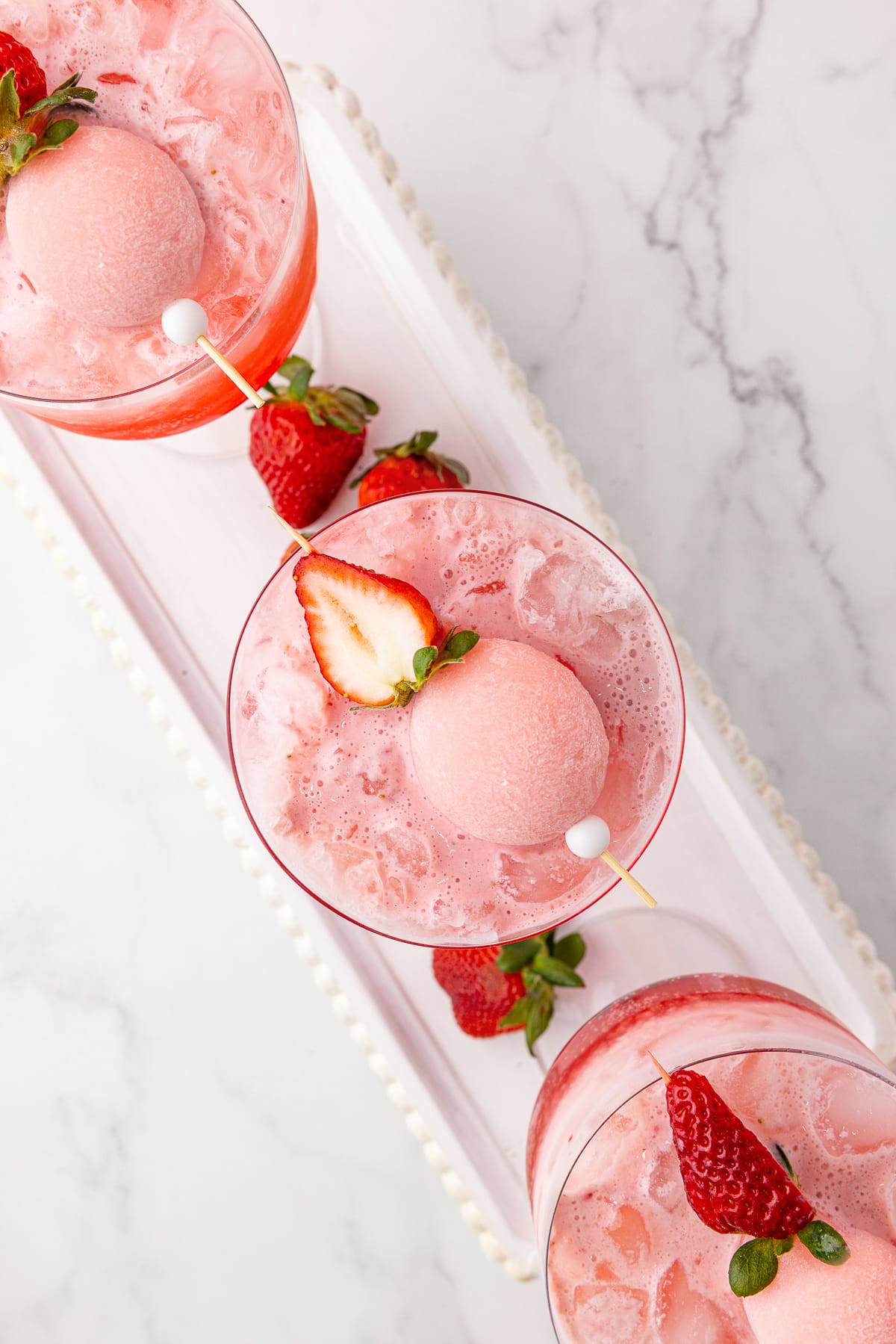 Overhead photo of pink drink with strawberry mochi on top of a coupe glass on a white tray with strawberries