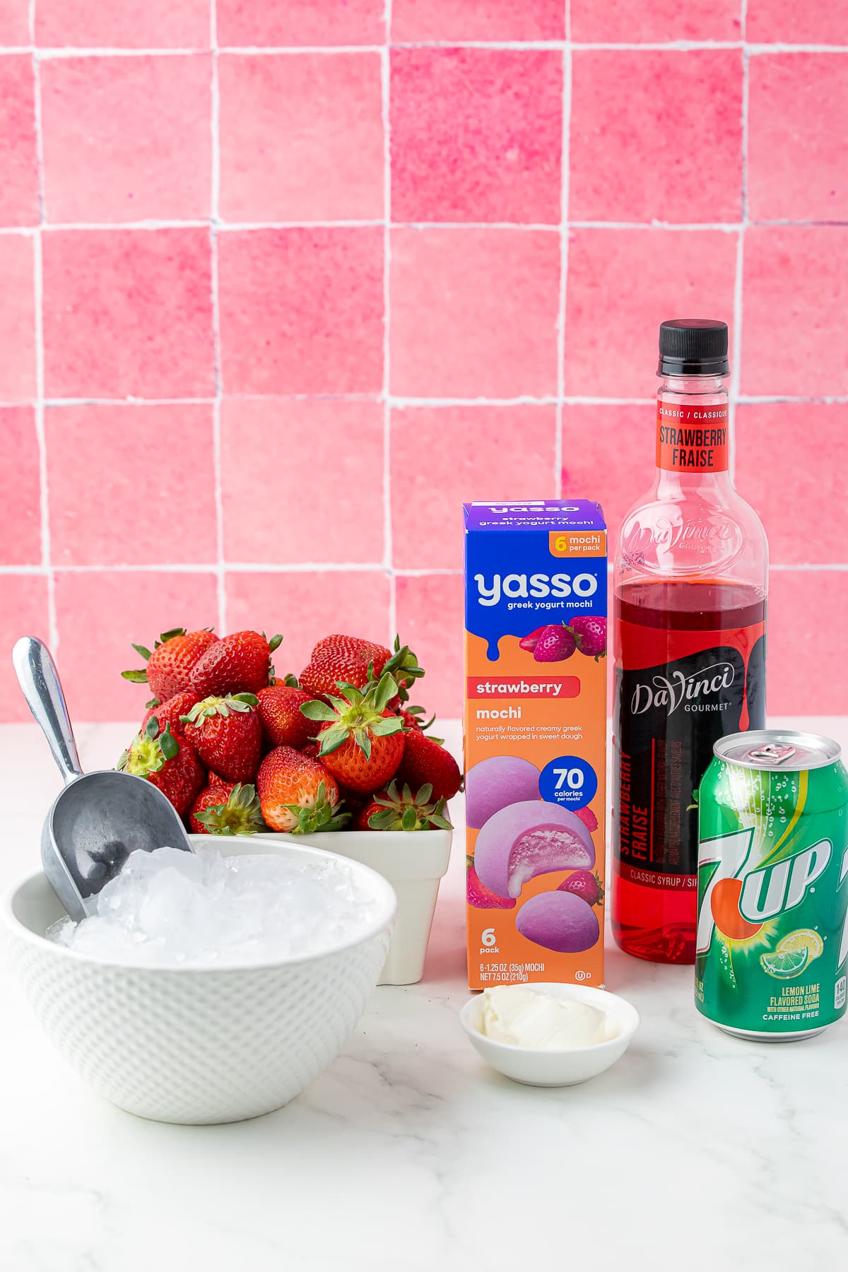 Ingredients for yassopink drink with strawberries, crushed ice, yasso mochi, strawberry syrup, 7up and greek yogurt on a white counter with pink backsplash