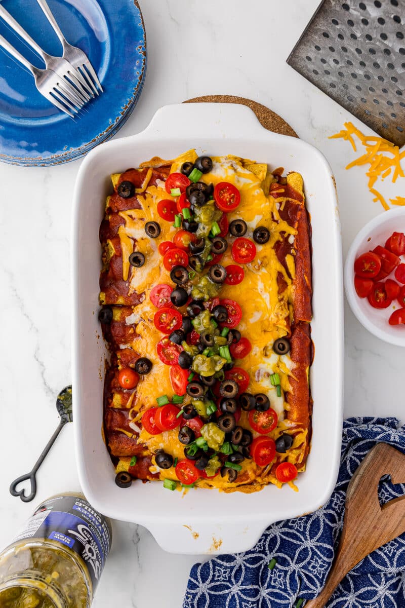 green onions, sliced olives, sliced tomatoes, on the taquitos casserole on a white countertop, with a blue dishcloth, heart spatula, and cheese grater