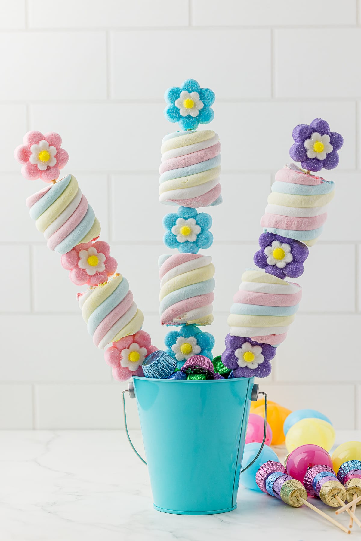 3 marshmallow candy kabobs in a blue pail on a white counter with more kabobs on the counter ready to insert into the candy bouquet