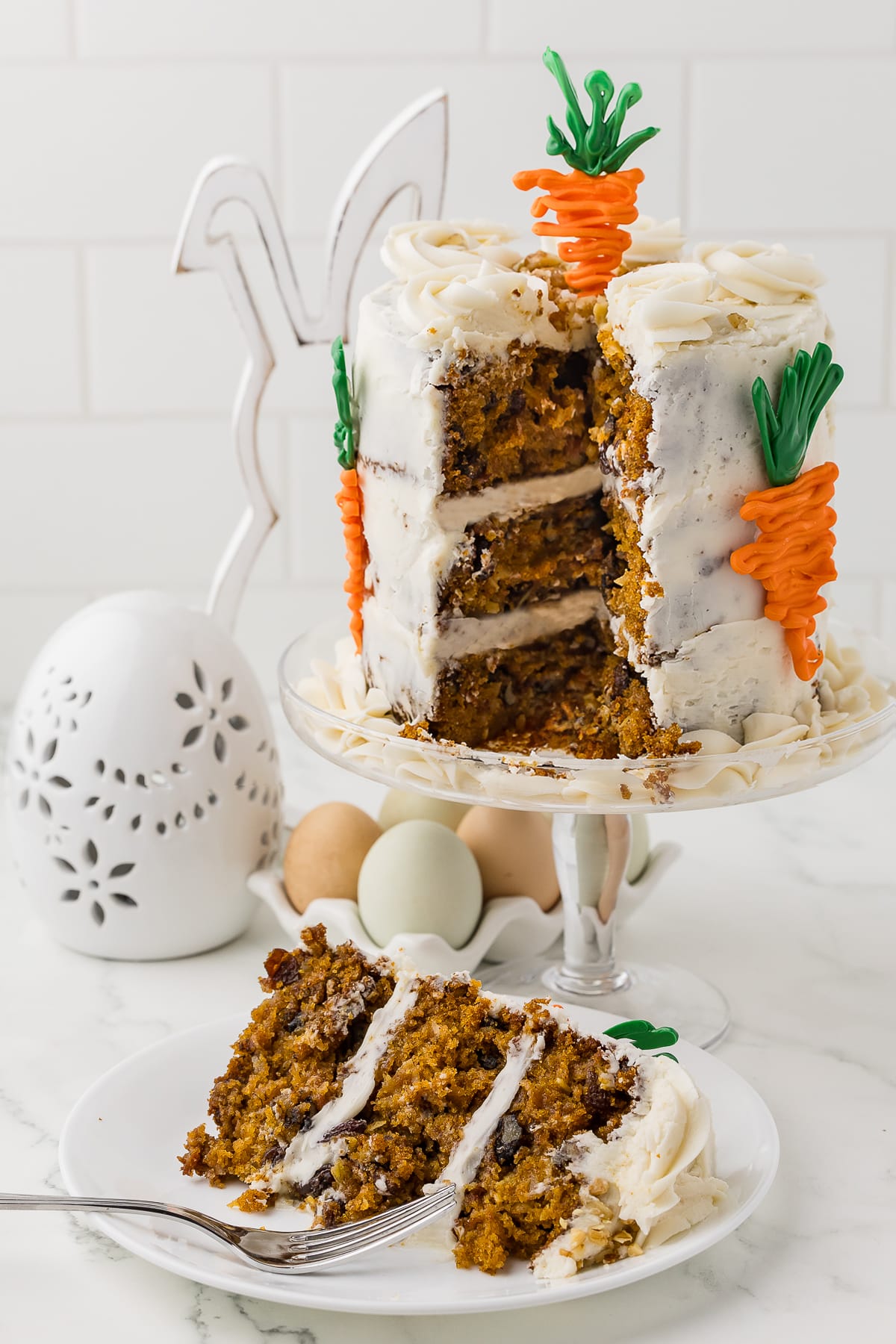 carrot cake with a slice taken out on a clear cake stand, white plate with slice of cake and a fork