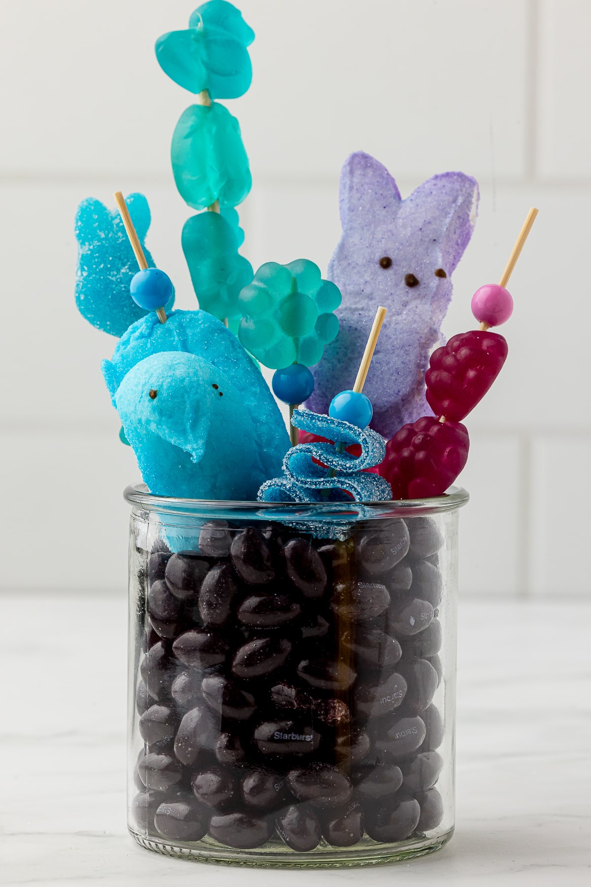 Purple jelly beans in a small jar with purple and blue candy kabobs made with blue gummy candy and purple candy bought at Five Below on a white countertop. Purple bunny peeps and blue chick peeps.