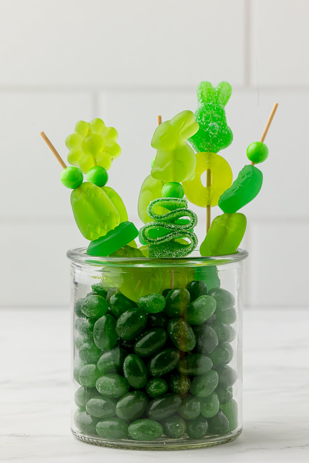 Green jelly beans in a small jar with green candy kabobs made with green gummy candy bought at Five Below on a white countertop.