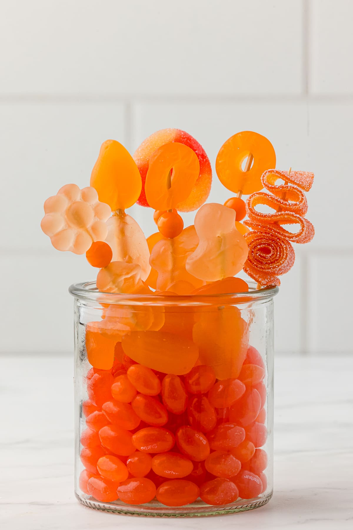 Orange jelly beans in a small jar with orange candy kabobs made with orange gummy candy bought at Five Below on a white countertop.