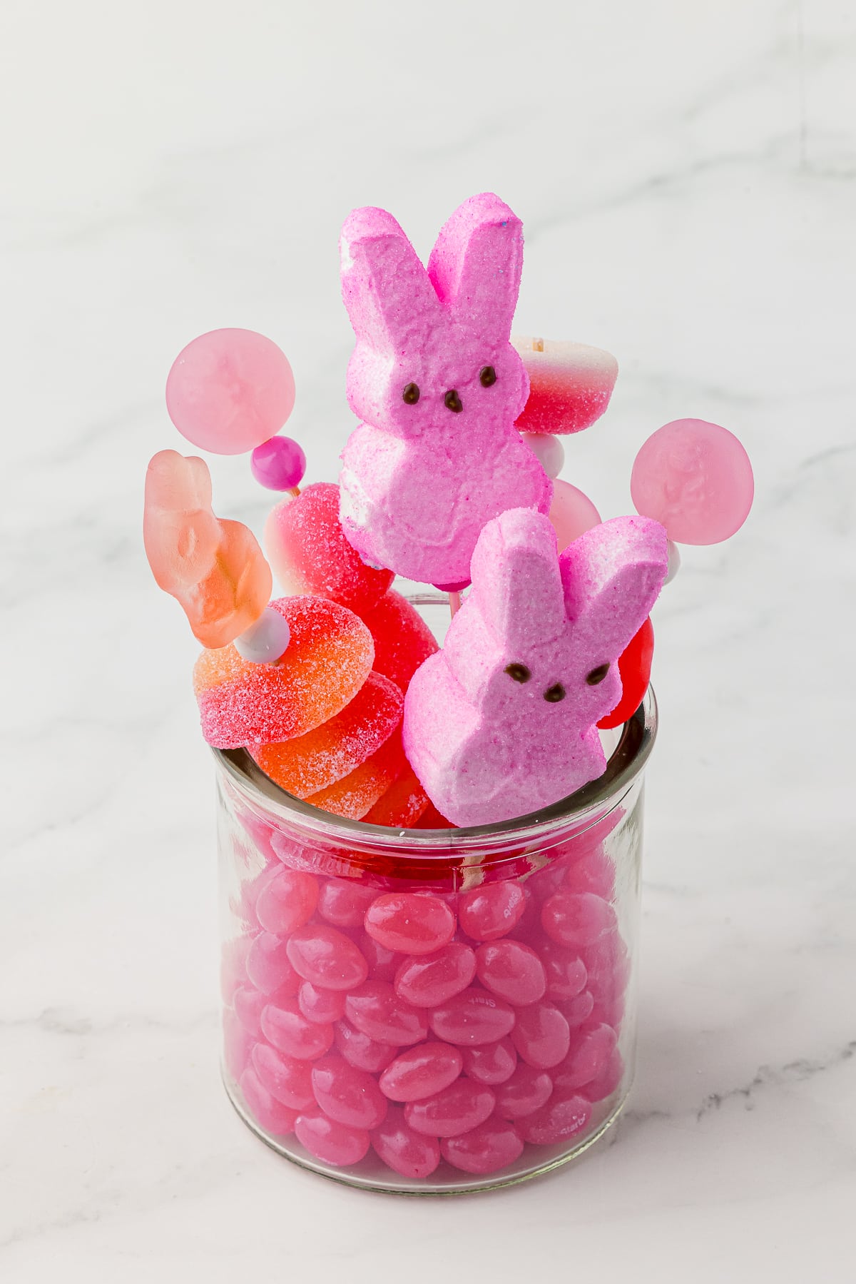 Pink jelly beans in a small jar with pink candy kabobs made with pink gummy candy and pink bunny peeps bought on a white countertop.