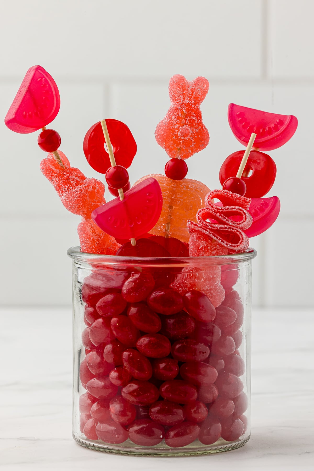 Red jelly beans in a small jar with red candy kabobs made with red gummy candy bought at Five Below on a white countertop.
