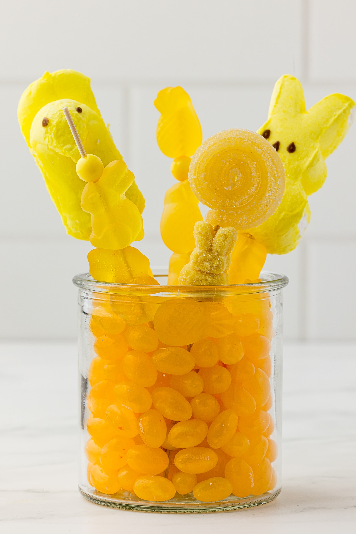 Yellow jelly beans in a small jar with yellow candy kabobs made with yellow gummy and yellow chick and bunny marshmallow peeps candy on a white countertop.