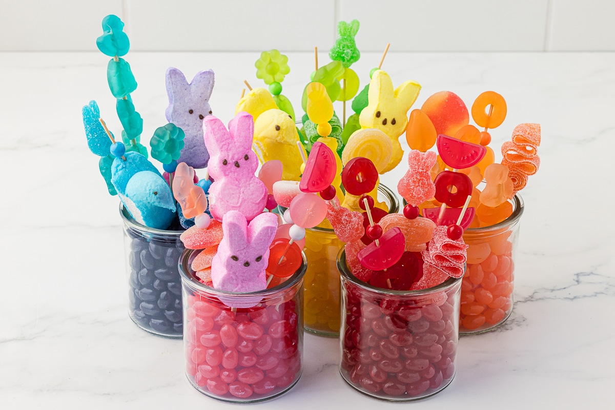 five jars filled with starburst jelly beans and bright Easter candy kabobs made of gummy candies and Easter bunny and chick marshmallow peeps. 