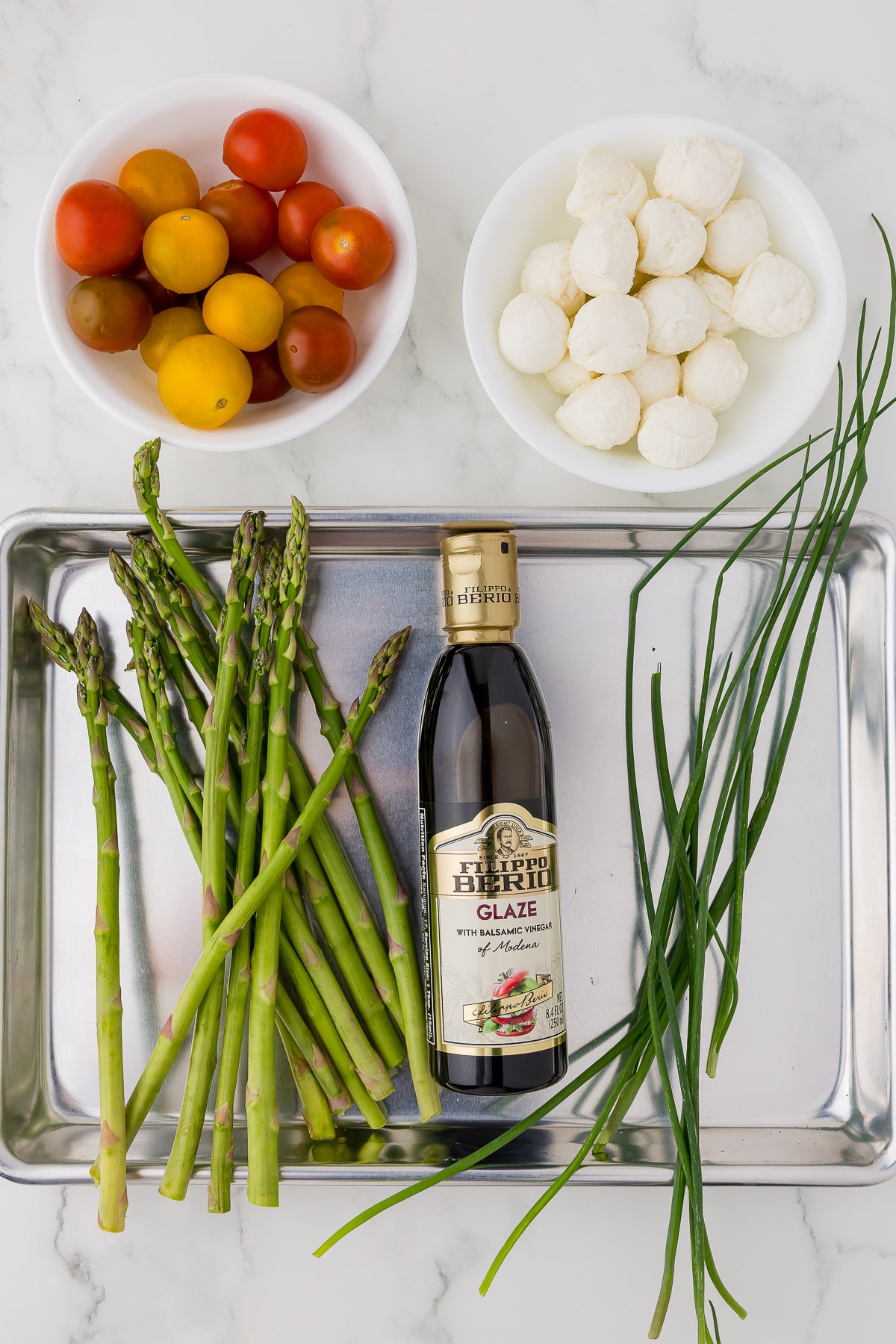 ingredients for tomato tulips: a bowl of yellow, variegated, and cherry tomatoes, a bowl of mozzarella balls, and a mini cookie sheet with asparagus spears, balsamic glaze, and chives