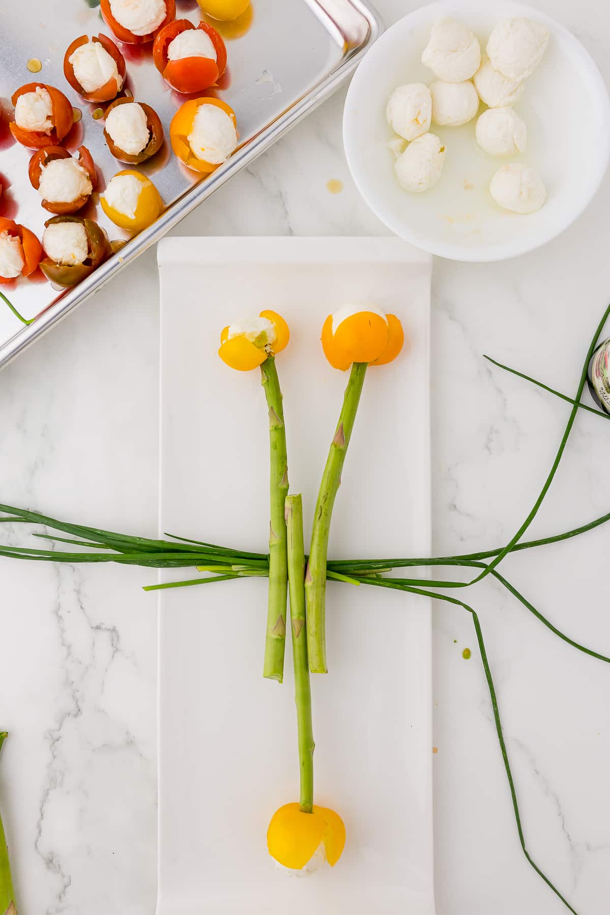 chives rope laying on a white platter with three tomato tulips on top with a white bowl with mozzarella balls and and a cookie sheet with tomato tulips ready to be speared with asparagus