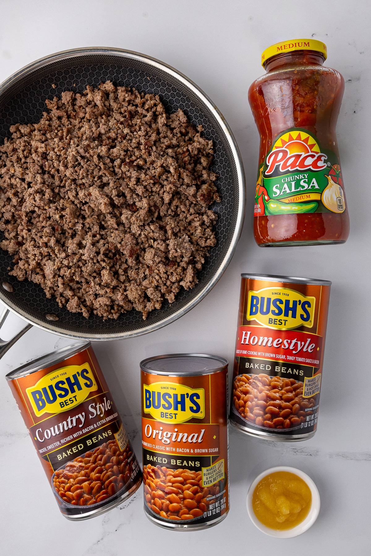 browned hamburger meat in a hexclad frying pan, a jar of pace chunky salsa, three cans of bush's best baked beans, and a small bowl of applesauce