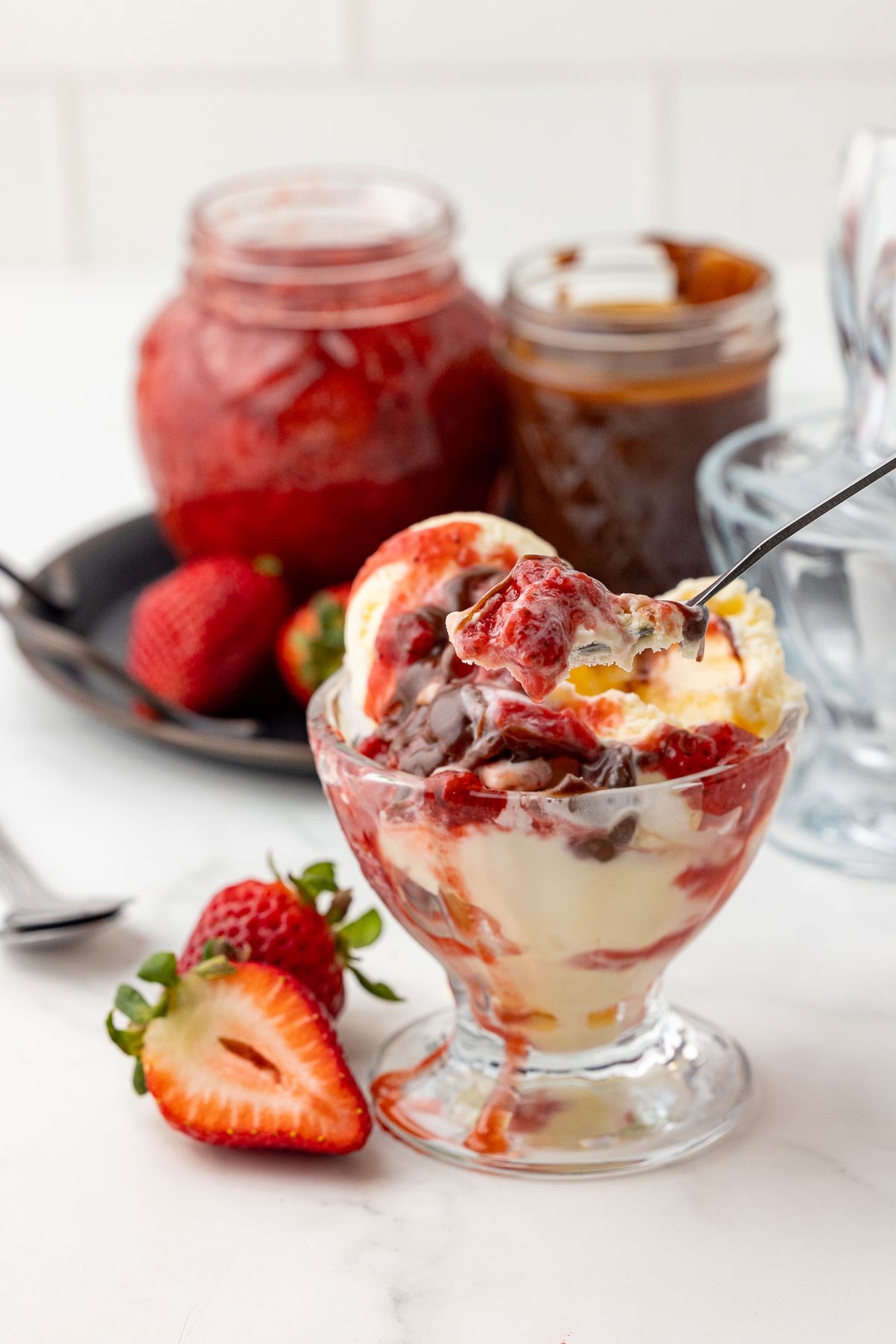 ice cream in a sundae cup topped with strawberry compote and chocolate sauce, with a jar of compote and a jar of sauce in the background