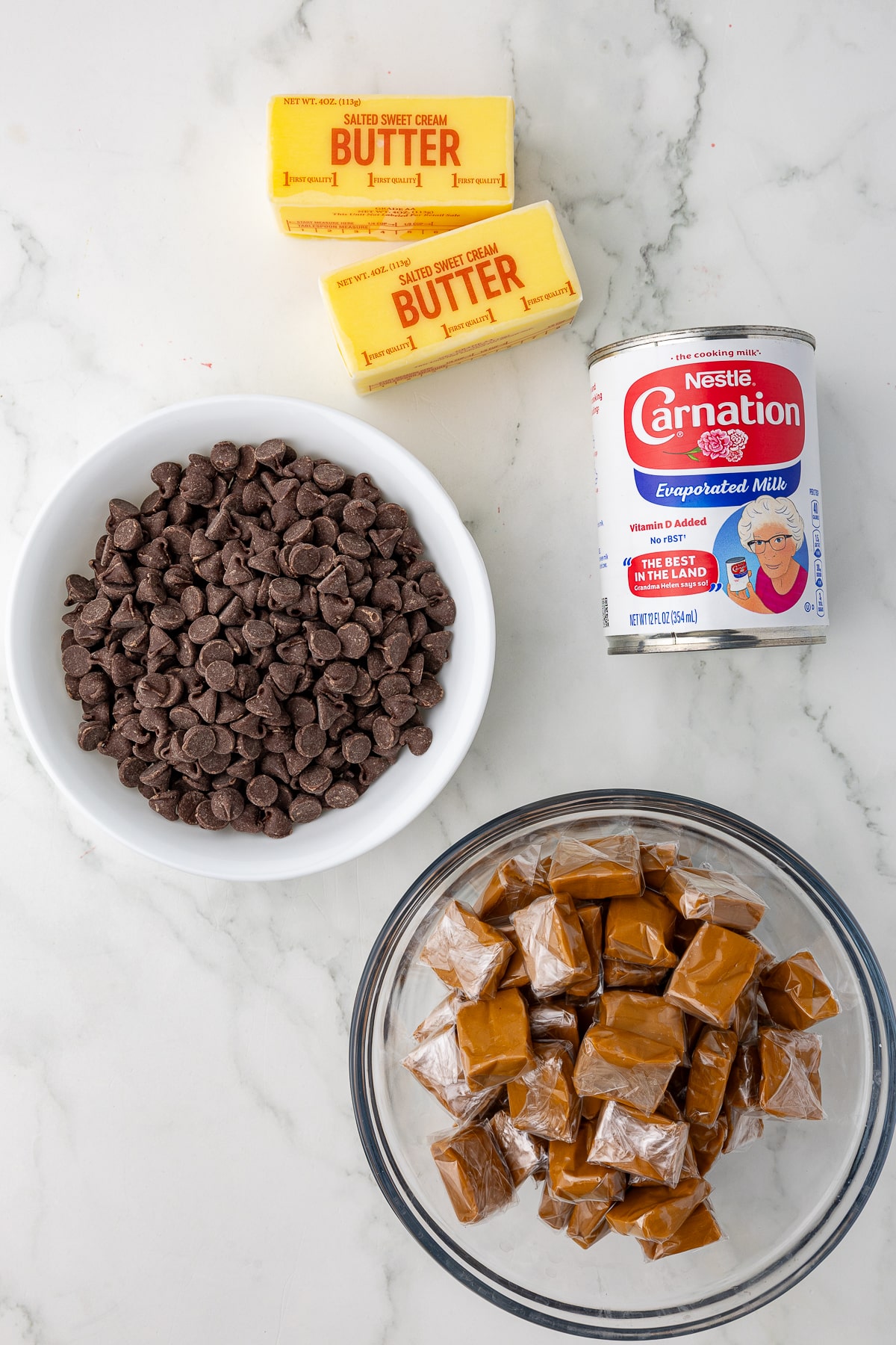 ingredients for chocolate sauce on a white counter, carnation evaporated milk, a stick of butter, a bowl of chocolate chips, and caramels in a glass bowl