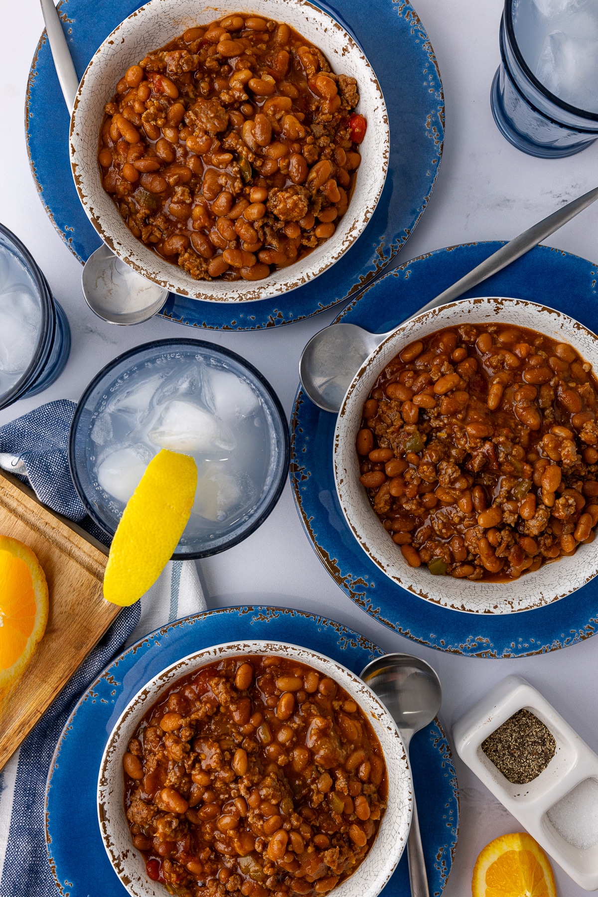 3 bowls of baked beans sitting on blue plates and a  striped napkin on a white countertop, with a soda, lemon wedge, and a salt and pepper holder