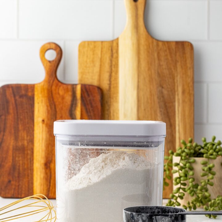 cake flour in an oxo container with a whisk, measuring spoons, a measuring cup, and two cutting boards on a white counter