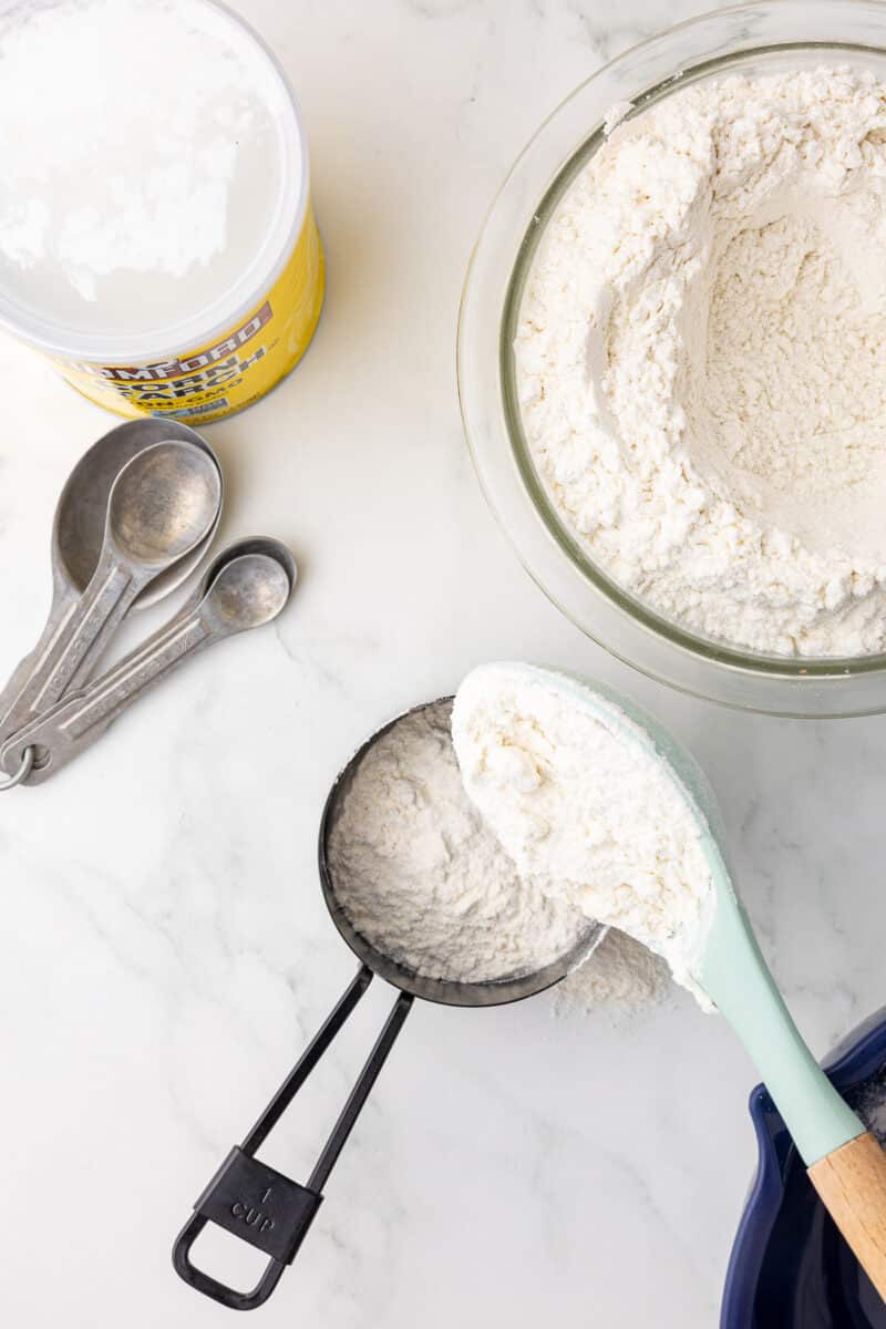 one container of cornstarch, measuring spoons, a black measuring cup with flour and a bowl of all purpose flour