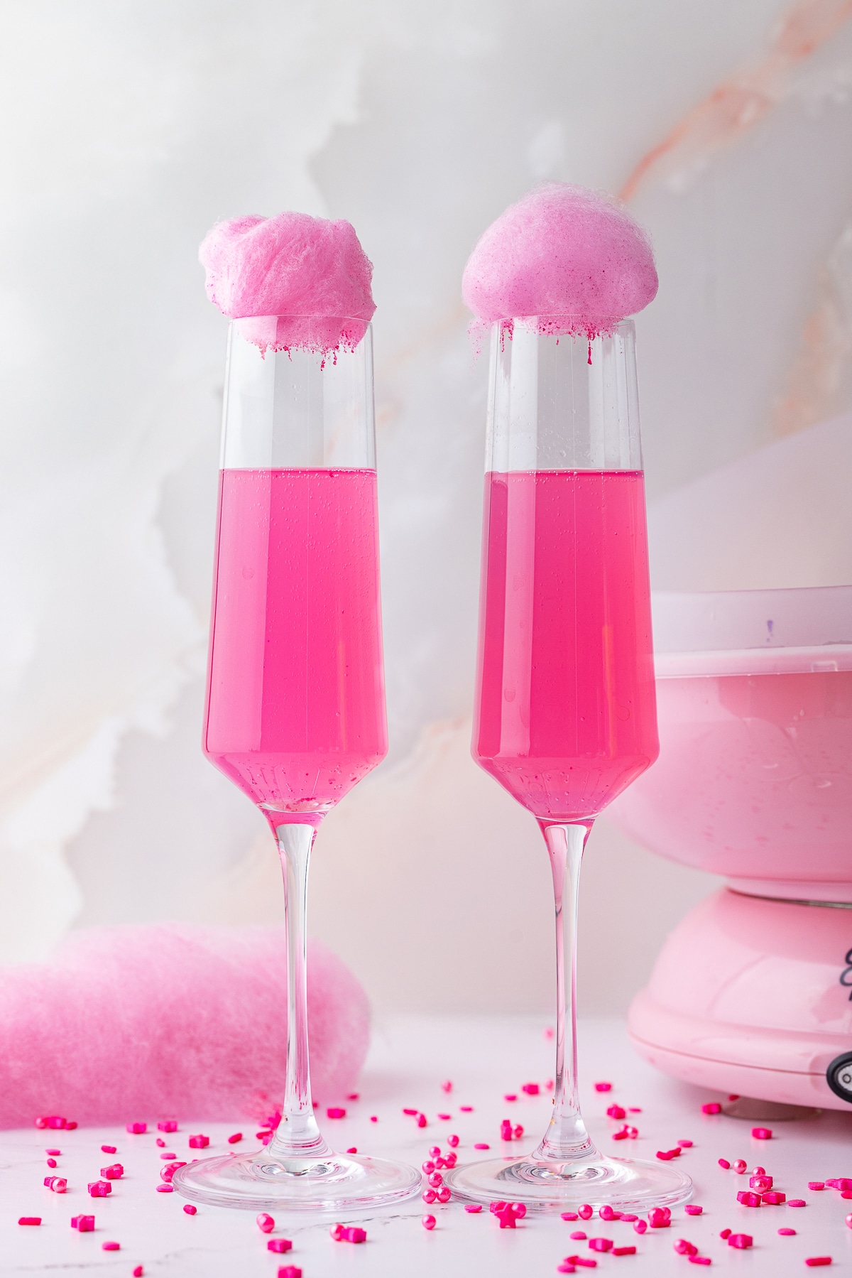 pink cotton candy over a 2 pink champagne flutes, Wilton sprinkles on a white counter with a pink cotton candy machine in the background. 
