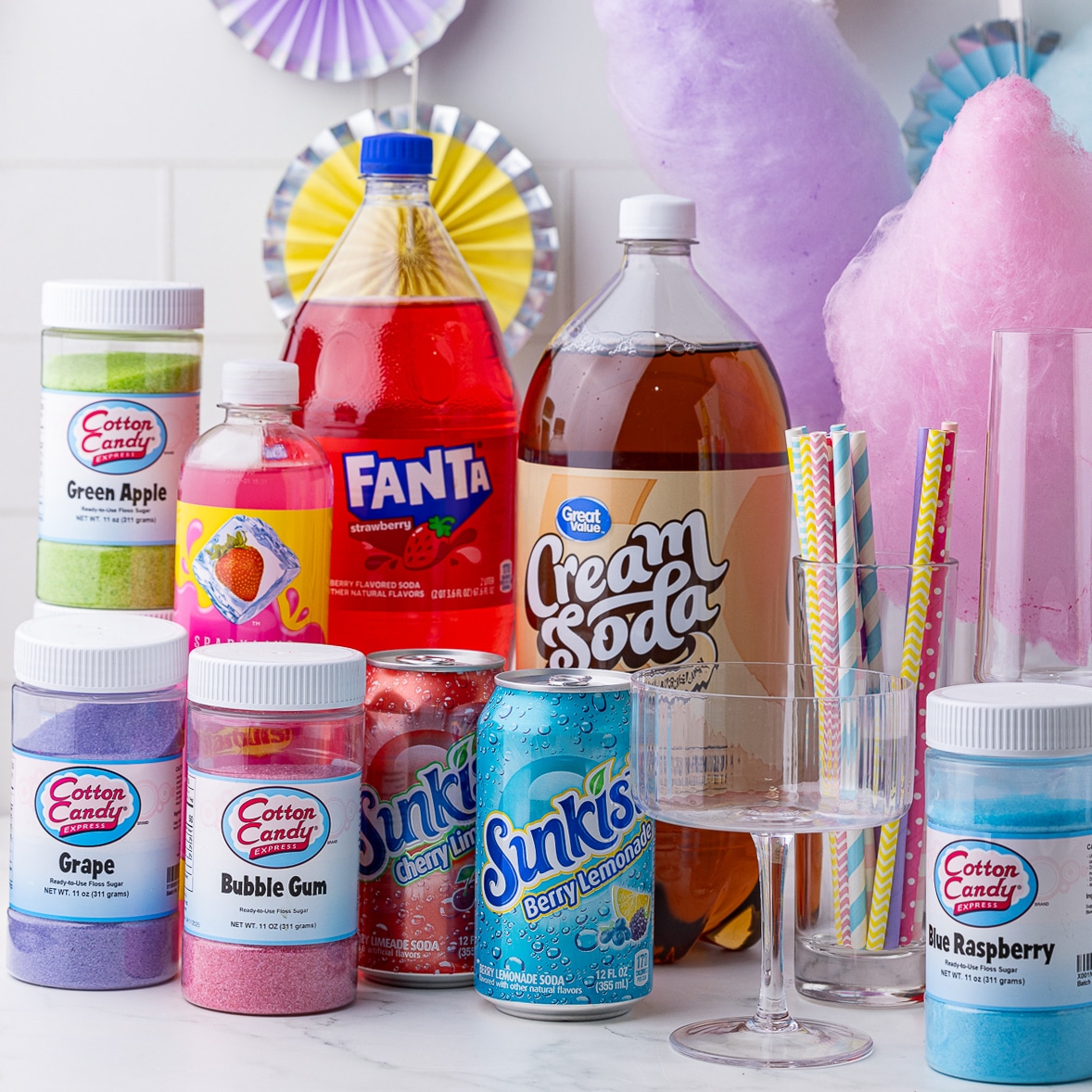 cotton candy sugar, assorted sodas, assorted drinking straws, and glasses on a white counter with purple, pink, and blue cotton candy in the background