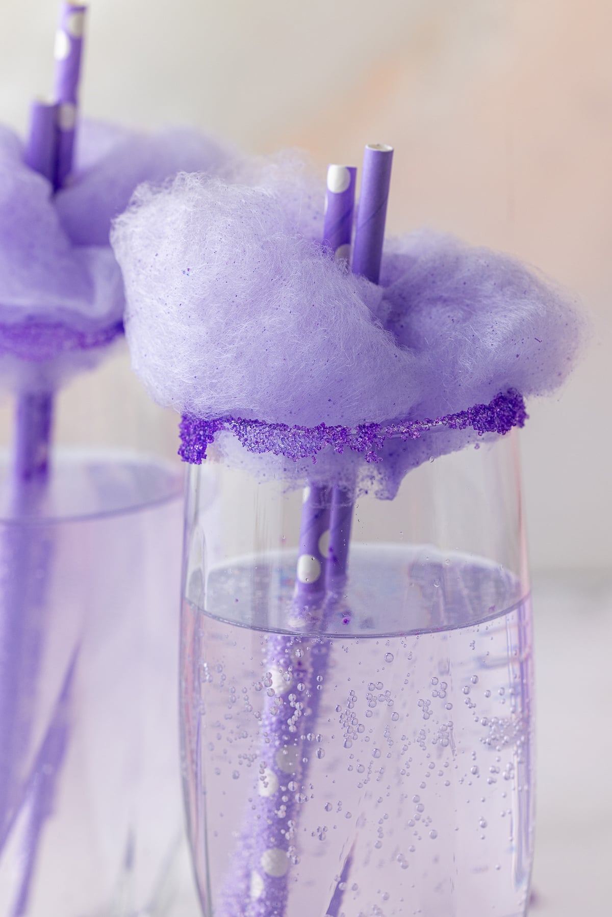 closeup view of purple soda in a glass topped with purple cotton candy, purple colored sugar on the rim of the glasses and purple straws