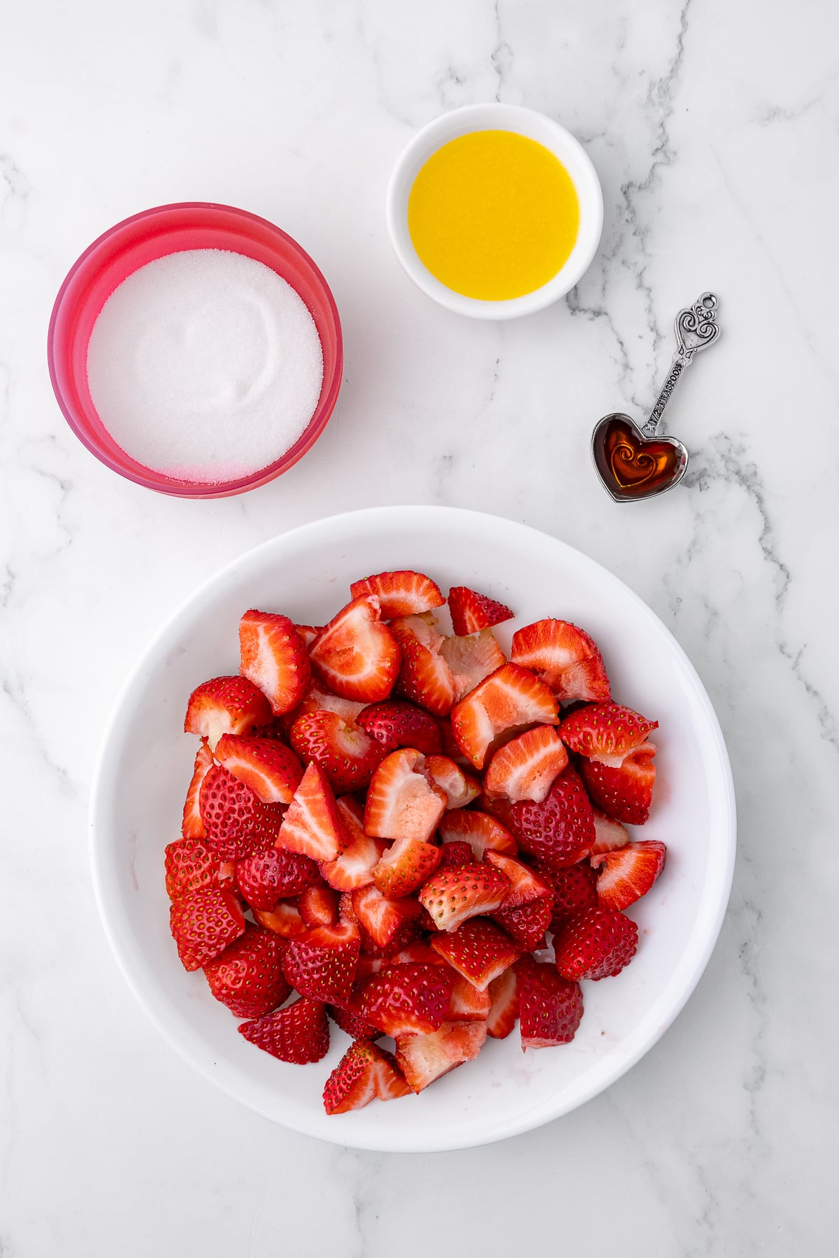 quartered strawberries in a white bowl, small bowl of sugar, small bowl of lemon juice, and a heart shaped teaspoon of vanilla