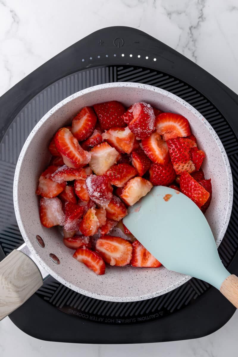saucepan with strawberries and sugar on a hotplate stirred with a blue spatula
