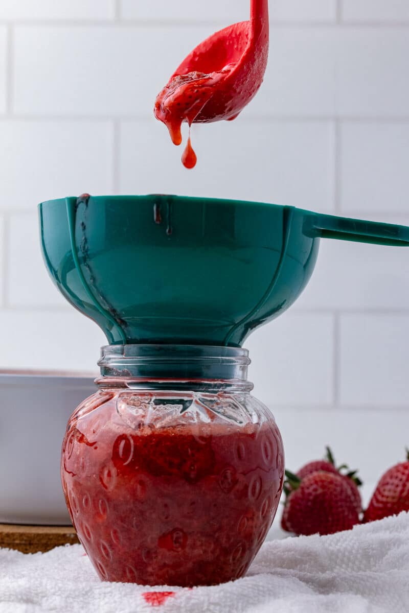 red ladle with strawberries pouring straberry compote into a green funnel and strawberry shaped glass jar