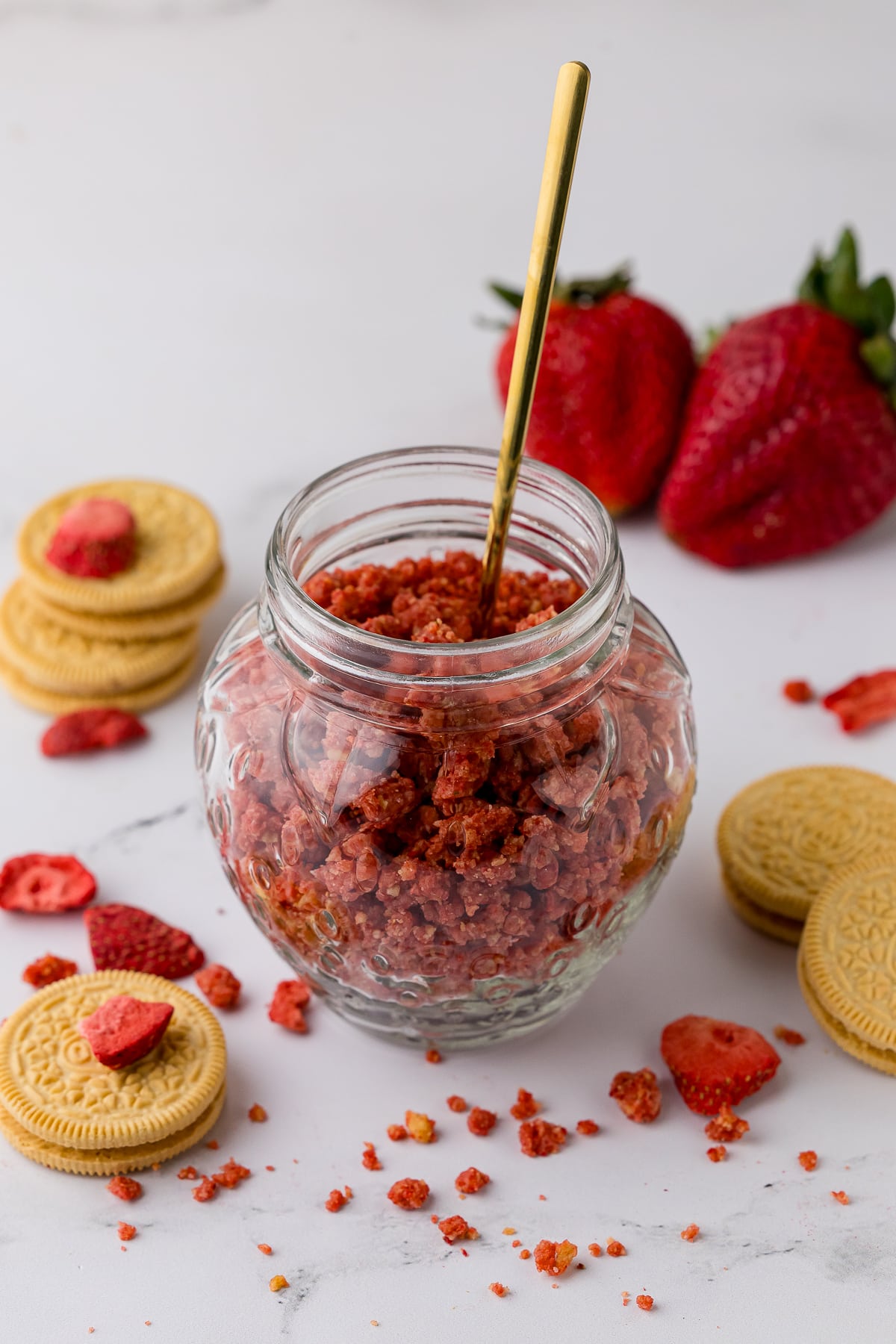 strawberry crunch in a glass strawberry shaped jar with a golden spoon inside and golden oreos, fresh strawberries, and freeze dried strawberries on a white countertop
