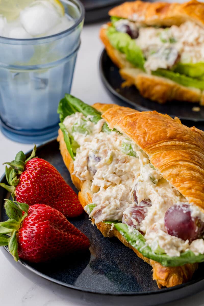 chicken salad croissant on a blue plate with two fresh strawberries with a blue glass filled with ice water and a lemon slice