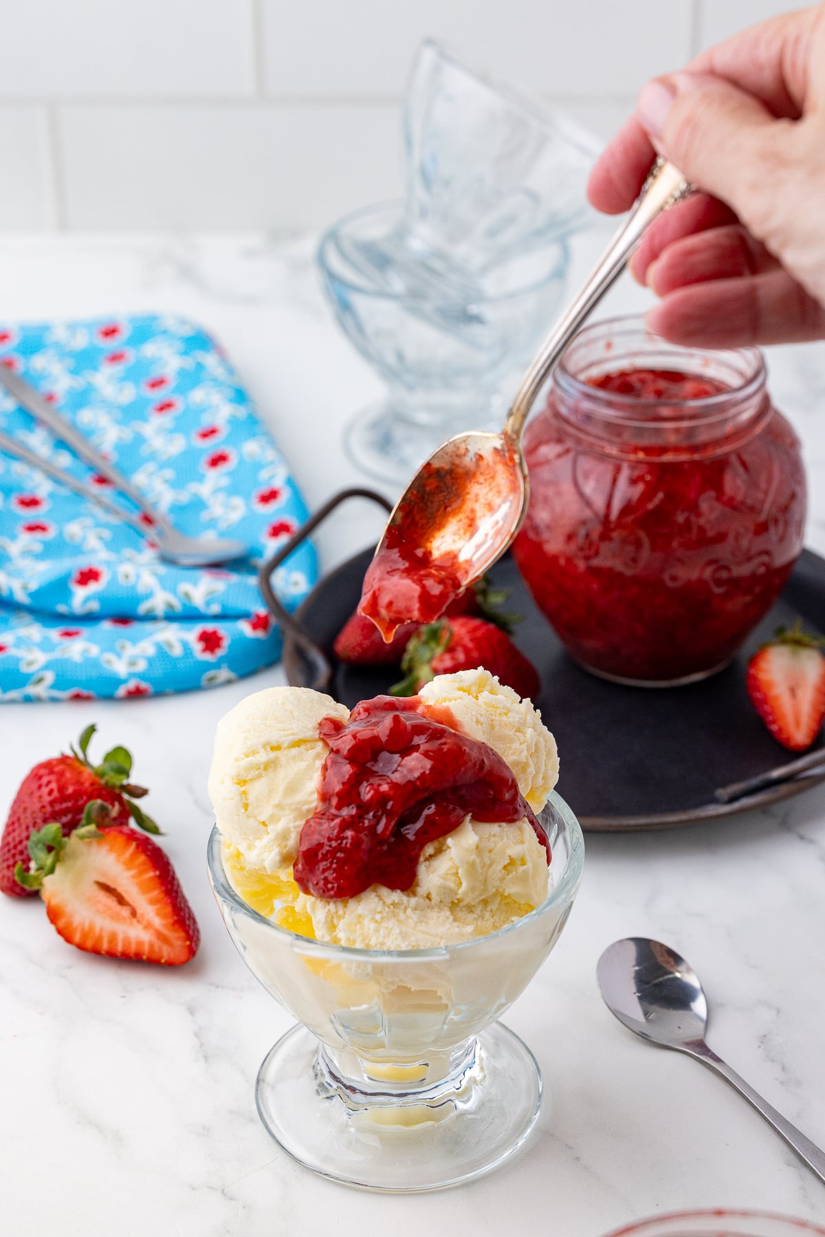 Strawberry compote spooned on three scoops of vanilla ice cream in an ice cream sundae dish with a strawberries, a blue dishcloth, and a strawberry shaped jar on a white countertop