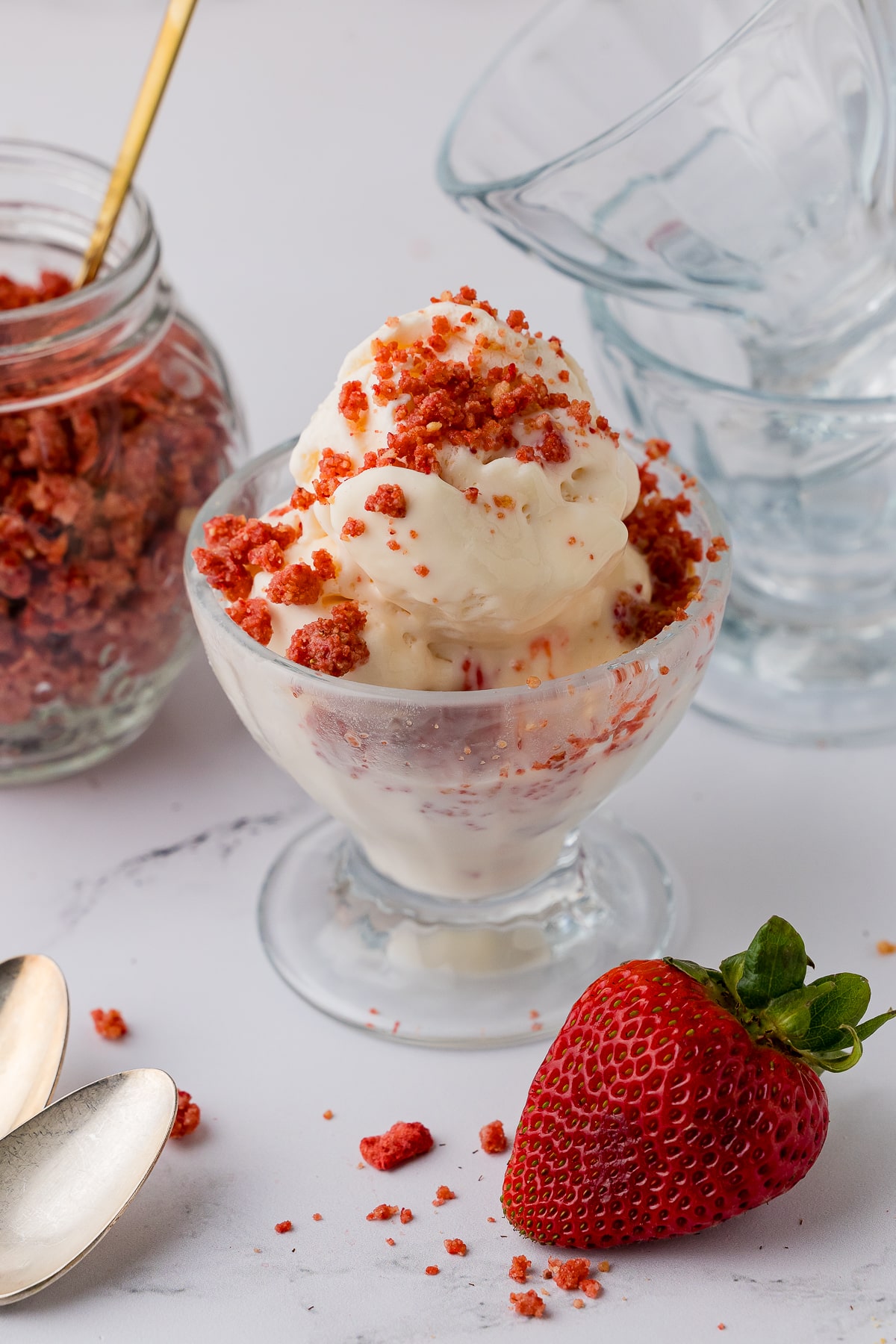 strawberry crunch sprinkled on a vanilla ice cream sunday with spoons, a fresh strawberry, extra glass sunday dishes and strawberry crunch on a white countertop