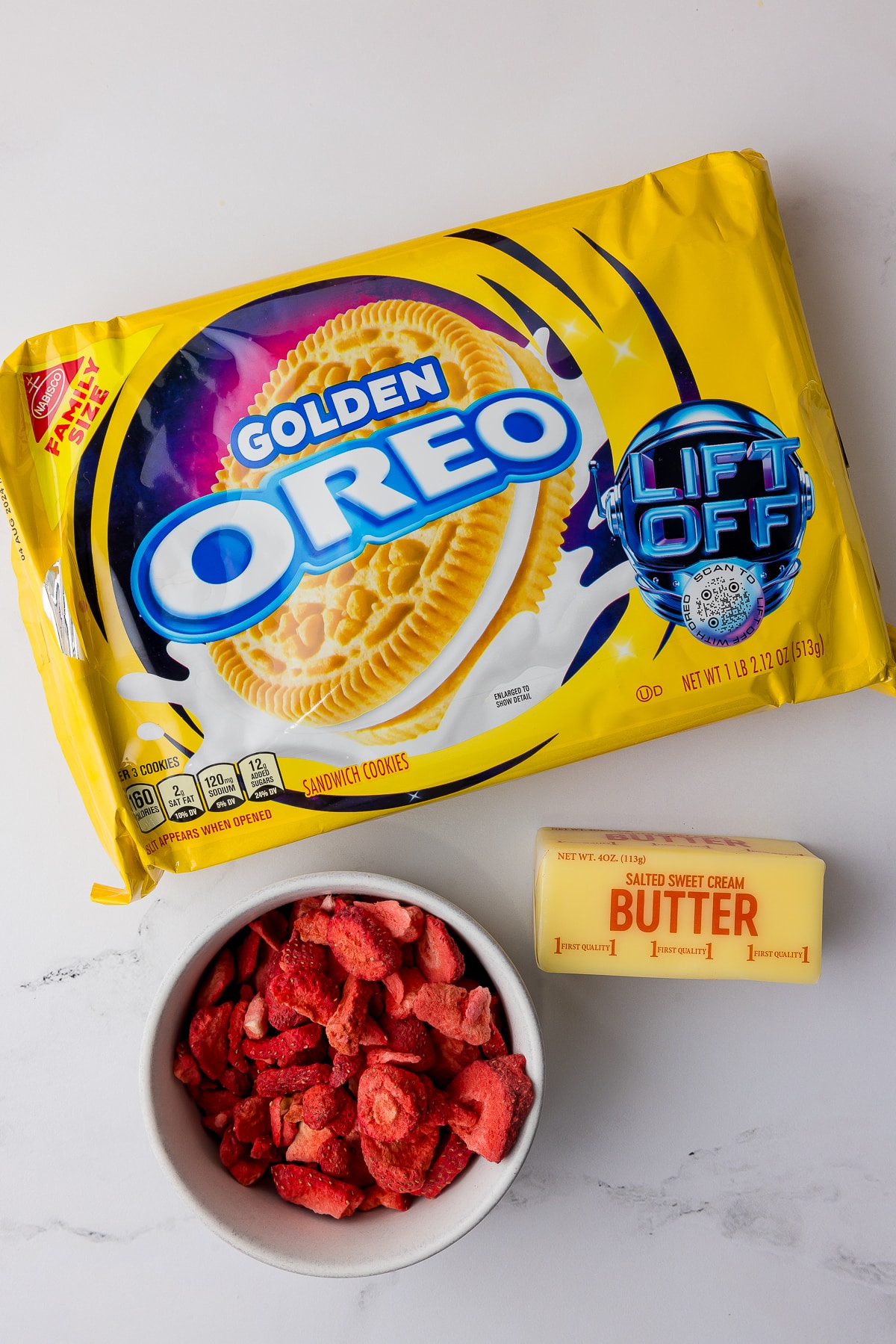 Package of golden oreos, white bowl of freeze dried strawberries, and a cube of butter on a white countertop