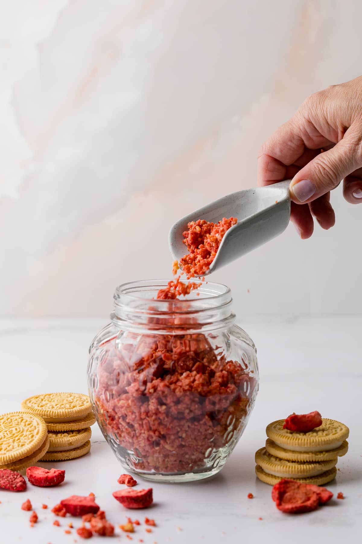 pouring strawberry crunch with a scoop into a strawberry glass jar and golden oreos and freeze dried strawberries on the counter