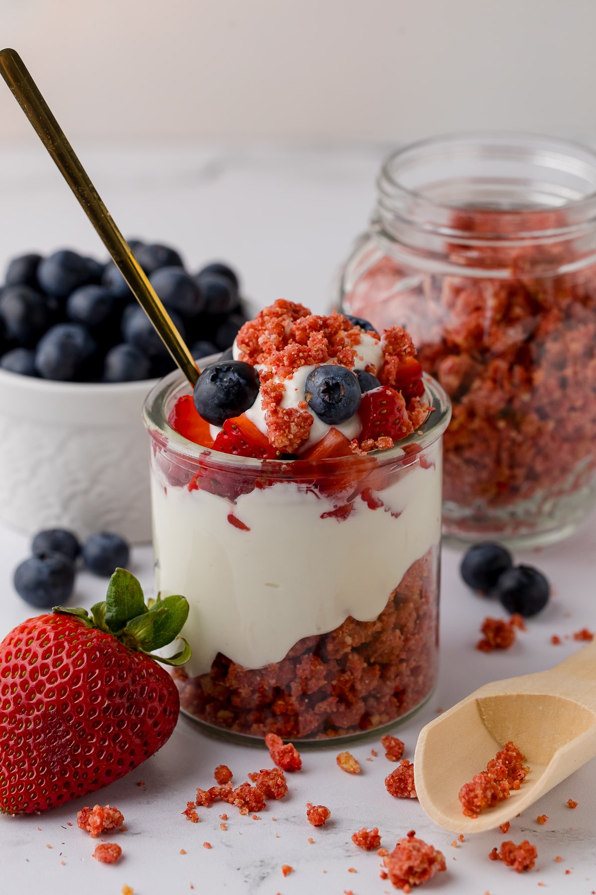yogurt and fruit parfait with strawberry crunch in the background, fresh strawberries, and a bowl of blueberries