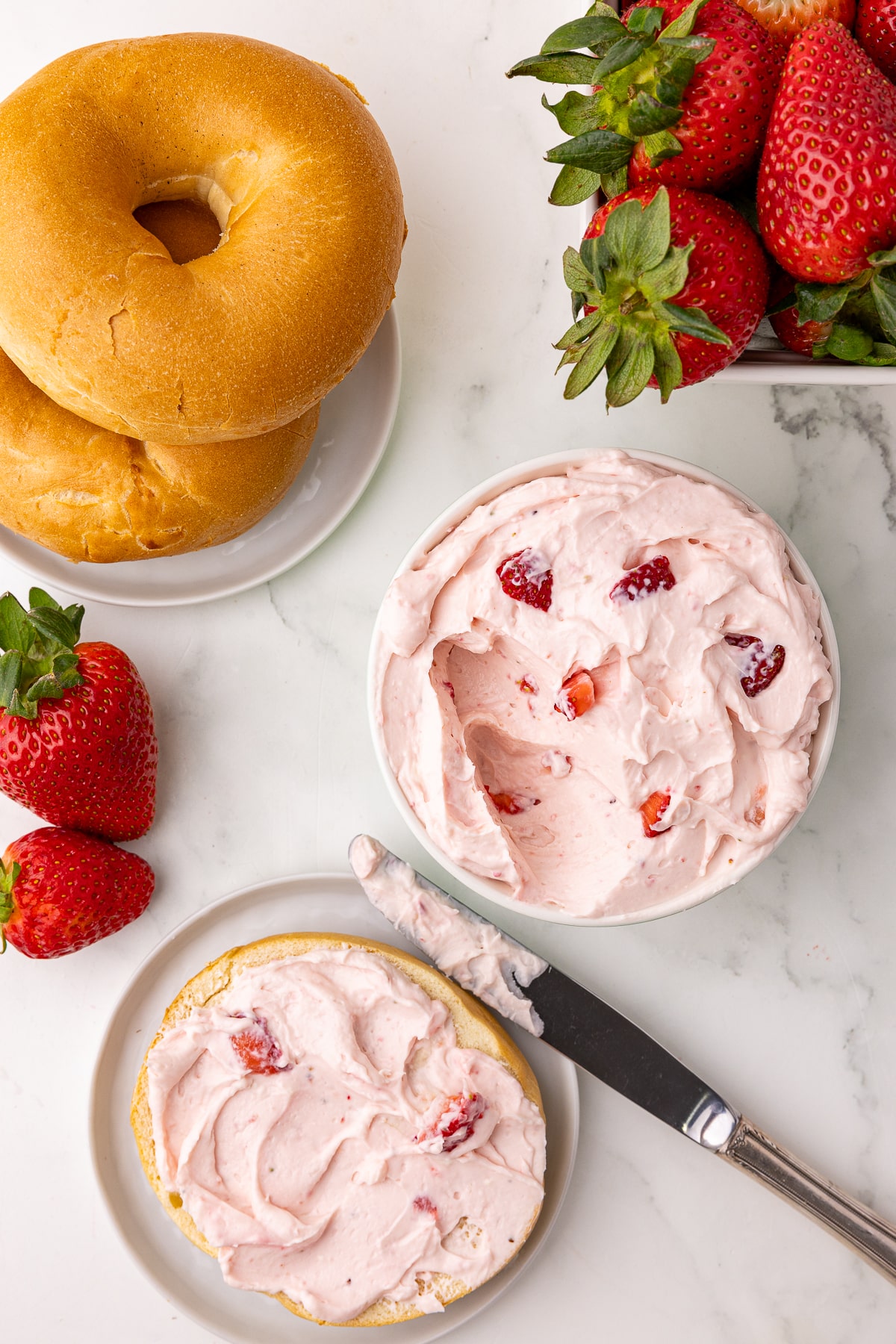 strawberry cream cheese on a plain bagel with a bowl of strawberry cream cheese, a berry basket with fresh strawberries