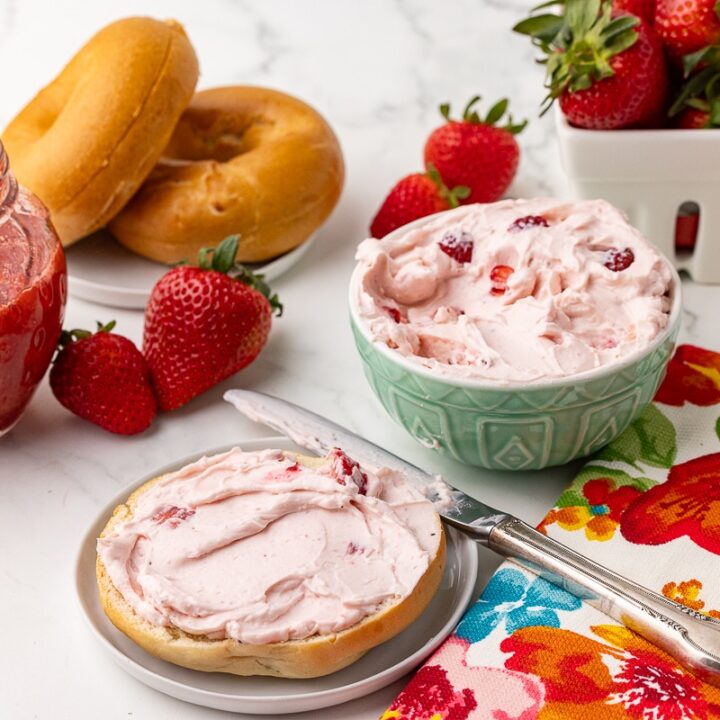 strawberry cream cheese on a plain bagel with a bowl of strawberry cream cheese, a berry basket with fresh strawberries, and a jar of strawberry compote