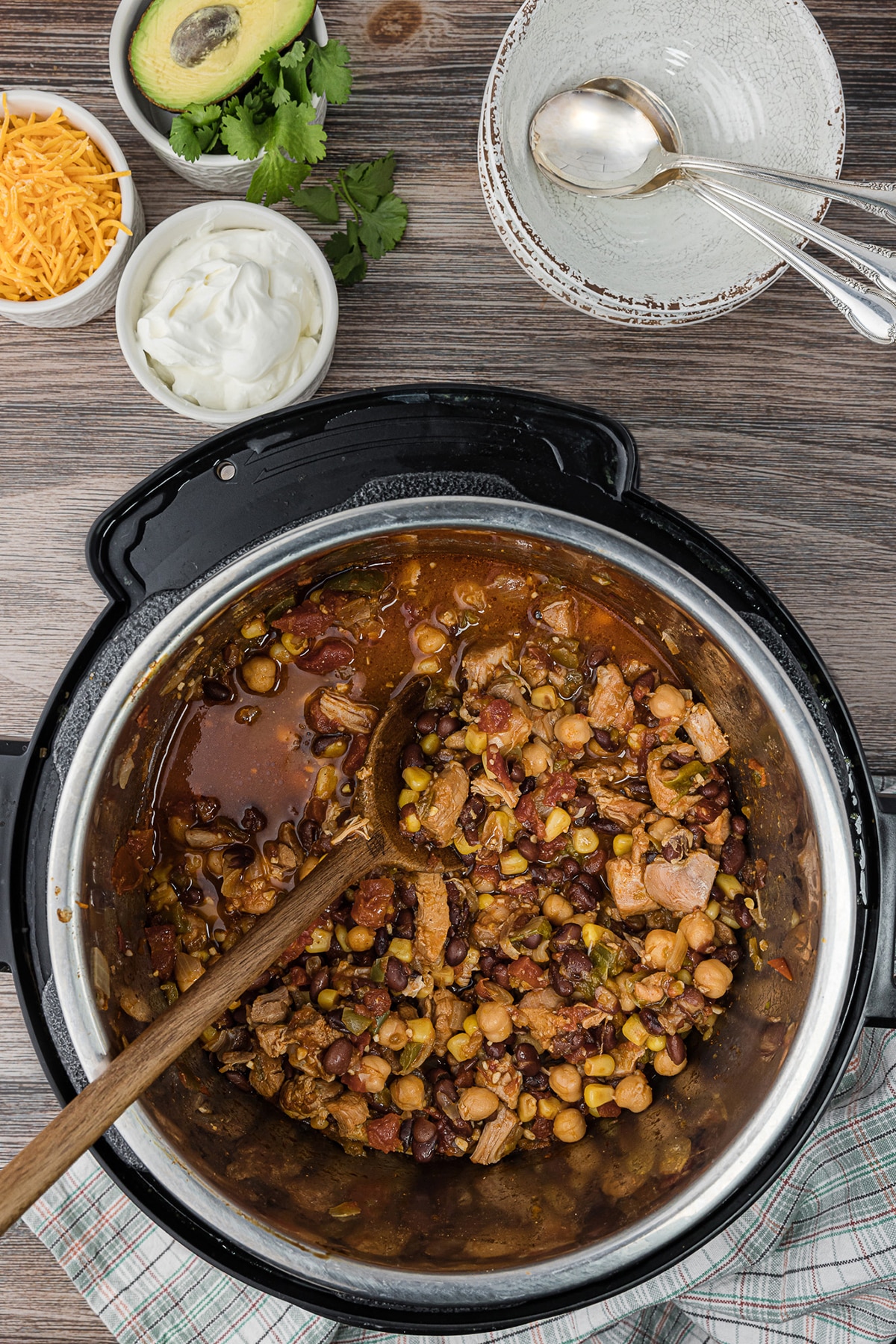 overhead photo of an instant pot of chicken chili on a wooden talbe with bowls and spoons, a small bowl of sour cream, a small bowl of shredded cheddar cheese, and a small bowl of avocado and cilantro